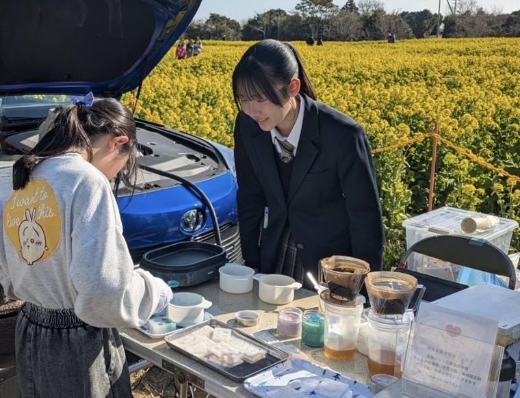 菜の花畑のそばで、ろうそくづくりの体験をする様子